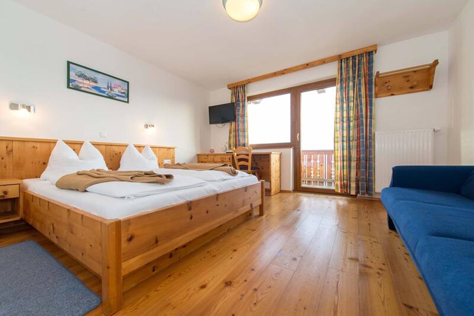 Gemütliches Hotelzimmer mit zwei Holzbetten, einer blauen Couch, einem Schreibtisch, einem Fernseher und großen Fenstern mit bunten Vorhängen.