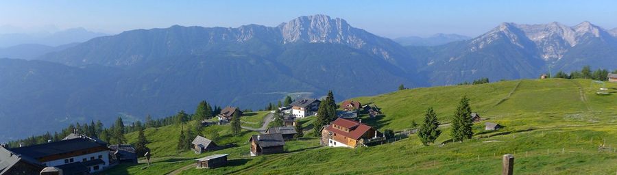 Eine malerische Alpenlandschaft mit sattgrünen Hügeln, charmanten Holzhäusern und hoch aufragenden Bergen unter einem klaren blauen Himmel.