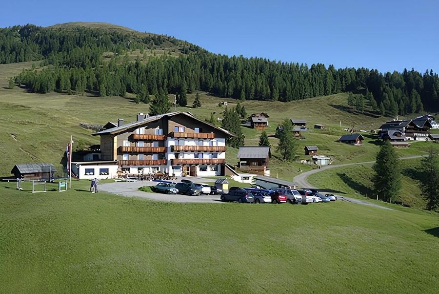 Malerische Alpenlandschaft mit einem Hotel im Chalet-Stil, umgeben von üppig grünen Hügeln und rustikalen Holzhäusern, mit geparkten Autos.
