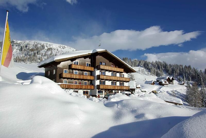 Charmantes Holzchalet, umgeben von tiefem, unberührtem Schnee, mit fernen Bergen und einer wehenden Fahne unter einem klaren blauen Himmel.