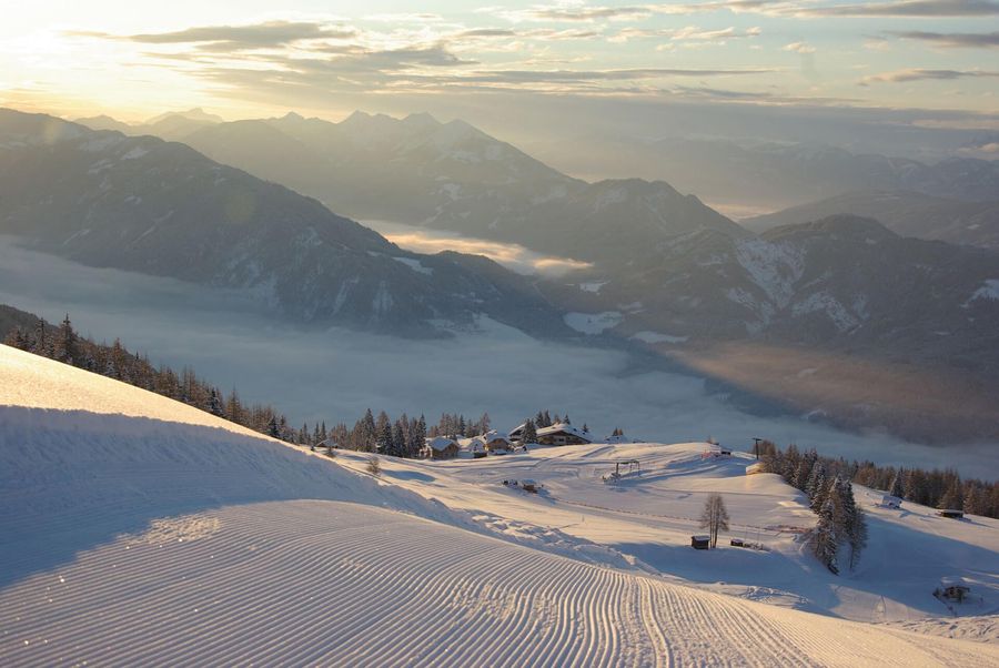 Schneebedeckte Berge erheben sich über einem nebelverhangenen Tal, das in goldenes Licht getaucht ist, und Skiloipen schlängeln sich durch die heitere Winterlandschaft.